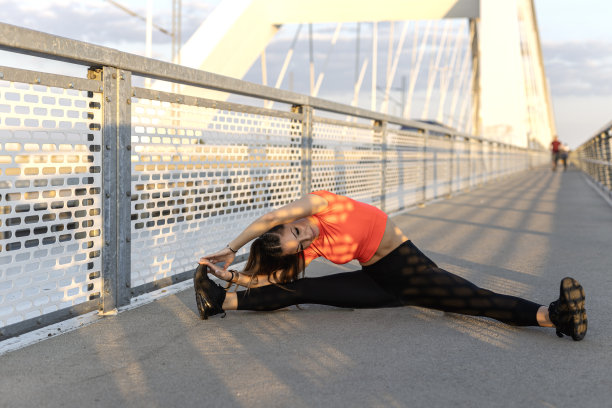 年轻快乐专注的健身女孩，穿着黑色瑜伽裤和橙色短裤，白天在桥上的人行道上锻炼和伸展身体。前视图。图片下载