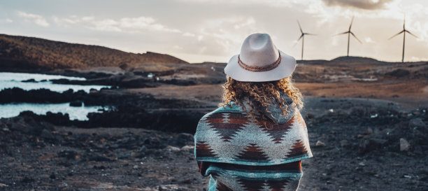 人和旅游目的地的概念生活方式。女人的后视图与时尚的帽子和夹克欣赏在背景风力磨坊在日落时间。可再生绿色能源生产图片下载