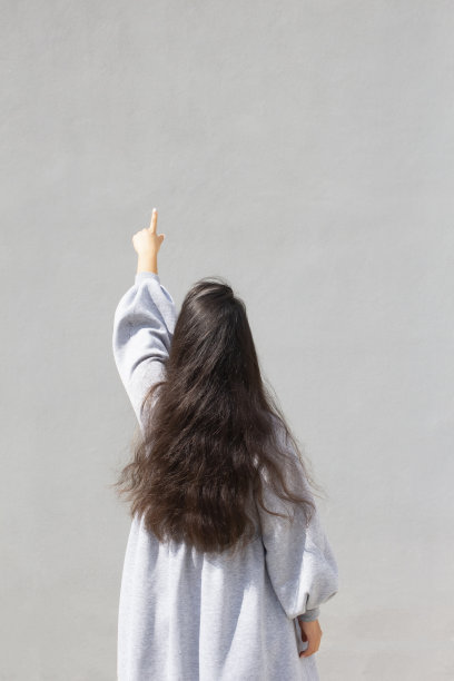 亚洲女孩长着黑色的长发，站在灰色的墙壁附近，用她的手指指着，后视图图片下载