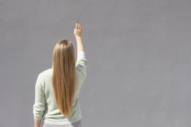 白种人的白色女孩长着自然的金色头发，站在灰色的墙壁附近，用她的手指指着，后视图图片下载
