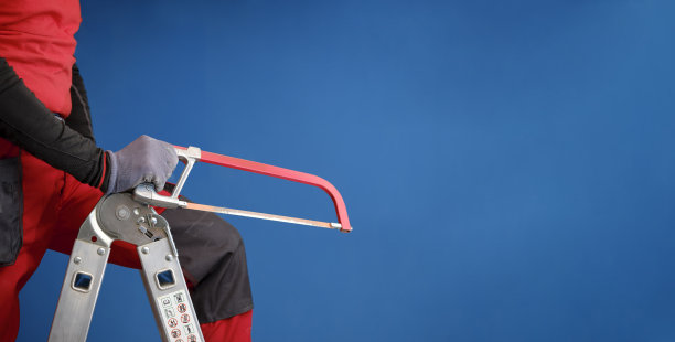 拿着锯子准备进行家庭装修的工匠。穿着工作服的工人在建筑工地的铝制折叠式梯子上。图片下载