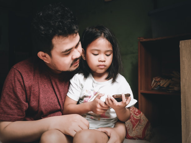 印度尼西亚雅加达，普拉加东- 2019年4月12日:一位父亲陪女儿玩智能手机图片下载