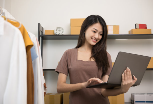 年轻美丽的亚洲女性商人在时尚商店持有笔记本电脑检查库存在轨道服装。在服装店工作的亚洲女性企业家。图片下载