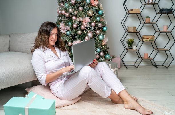 快乐的迷人的黑发女人使用笔记本电脑坐在沙发附近的地板上，工作和使用笔记本电脑在圣诞节图片下载