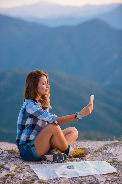 日落时分，一名年轻女子一边用手机自拍，一边惊叹着从世界之巅俯瞰的美景。成功，赢家，禅，宁静。图片下载