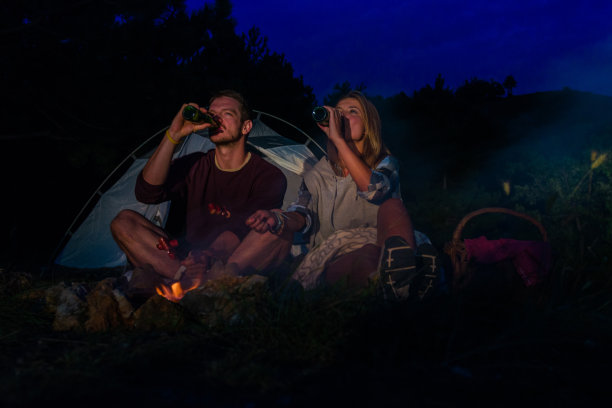 年轻的夫妇在篝火上烤香肠，在黄昏的森林山喝啤酒图片下载