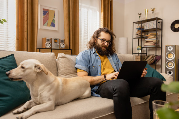 一位满脸笑容、戴着眼镜的大胡子男子在他心爱的狗狗陪伴下远程工作。这个人用他的笔记本电脑，浏览他喜欢的网站，给同事发电子邮件图片下载