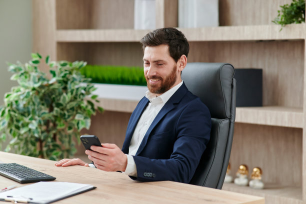 商人微笑着在办公室用他的智能手机输入信息。他穿着西装，工作场所是在现代和漂亮的办公室图片下载