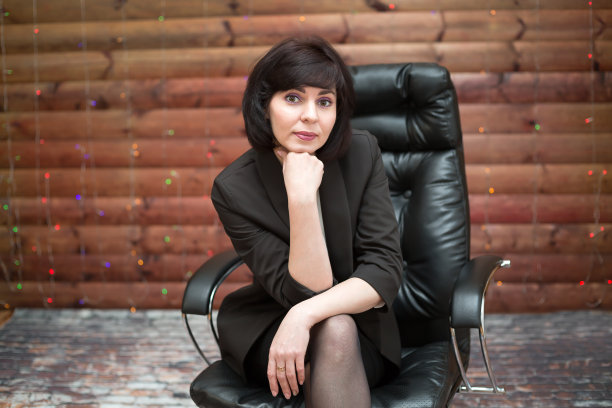 一位棕色头发的成年女性穿着黑色正装夹克，坐在木制背景的办公椅上图片下载