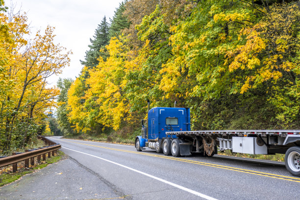 商业经典的蓝色大型拖车半挂车与空的平板半挂车运行在蜿蜒的山路上，黄色的秋天树木在斜坡上图片下载