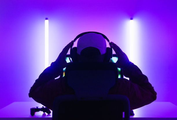 后视镜年轻播放器视频电脑pc。一个年轻人坐在游戏站的椅子上。一名男子戴着耳机在暗房里玩在线动作游戏。图片下载