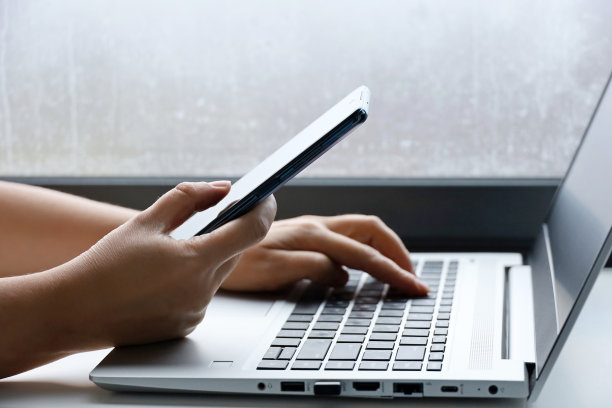 一个女人拿着智能手机坐在窗边的笔记本电脑键盘前图片下载