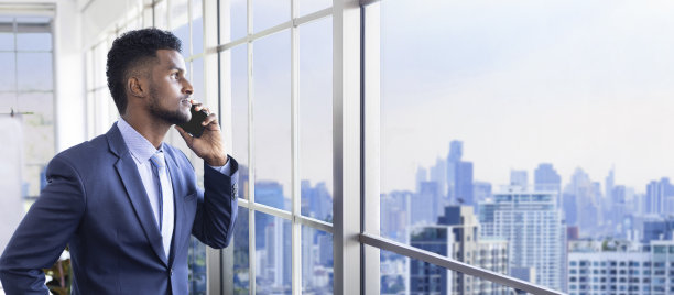 身着正装的非洲裔美国商人一边用手机看窗外的城市风景，一边眺望着摩天大楼的远景、梦想和成功的商业和投资规划图片下载