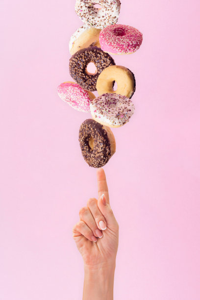 美丽的女人的手触摸坠落的环形甜甜圈与白色，巧克力和粉红色釉和彩色碎屑在粉红色的粉彩背景。图片下载