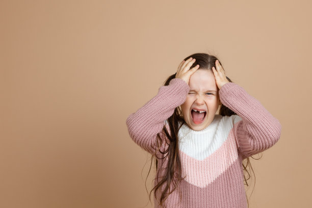 年轻愤怒的女孩的肖像，长着黑色的长发，穿着粉红色，白色的毛衣，张开嘴站着，呼喊着，用手托着头，在棕色的背景上显示出恶意。图片下载