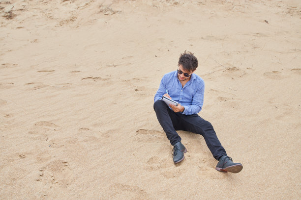 一个白人男子穿着正装坐在沙滩上写笔记本。图片下载