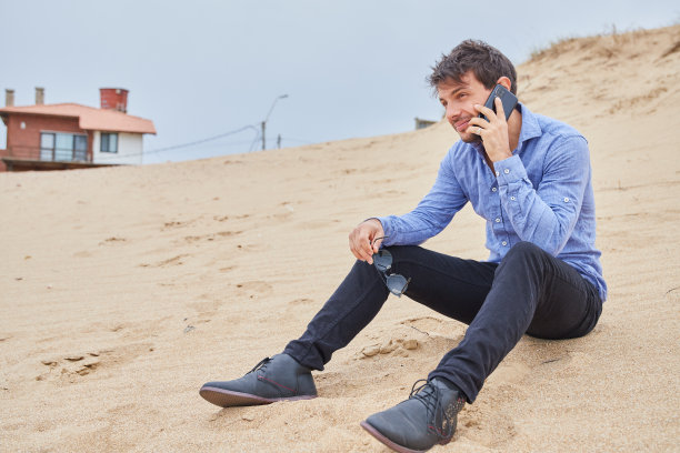 一个白人男性手里拿着墨镜在用智能手机聊天。他穿着浅蓝色的衬衫，黑色的裤子和黑色的鞋子。图片下载