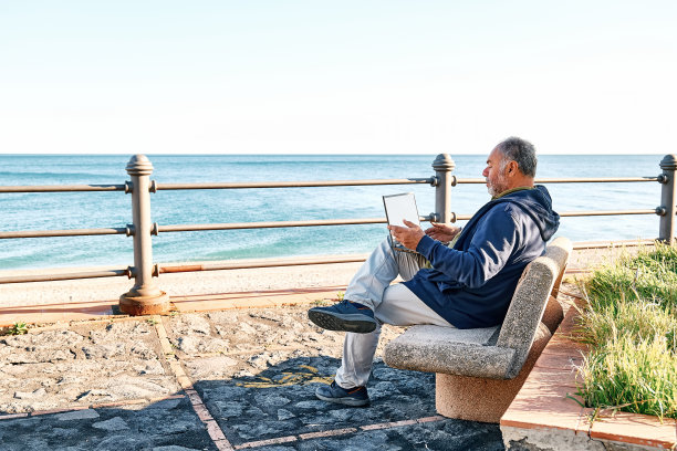 英俊的大胡子成熟男人，坐在海边的长椅上，带着笔记本电脑在户外工作。现代生活方式，联系，商业，自由职业的概念。图片下载