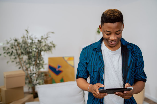 小伙子面带微笑地站在公寓中央，谈论着装修，新房子，搬箱子站在客厅里，男孩手里拿着平板电脑，在网上买公寓家具图片下载