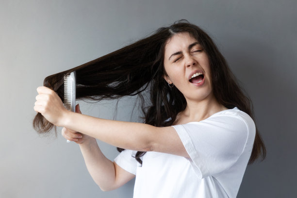 一名白人年轻女子用一把刷子梳理她纠结的黑发的肖像。灰色的背景。护发的概念图片下载