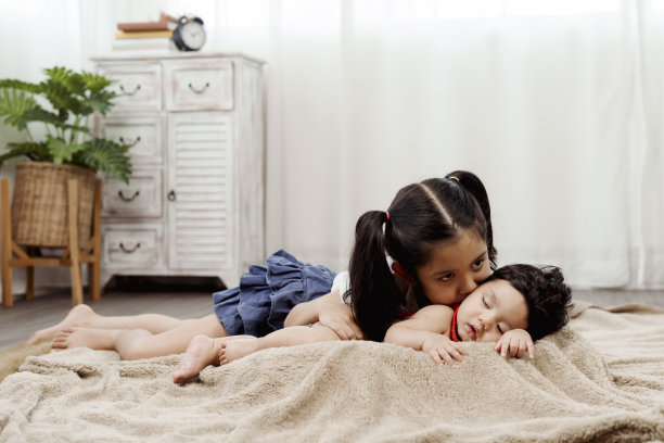 在客厅的地毯上，一个可爱的亚洲女孩抱着她刚出生两个月的弟弟，用爱和温暖亲吻弟弟的脸颊。幸福的家庭概念图片下载