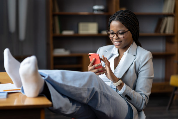 快乐的非洲女性坐在办公桌前用智能手机浏览社交媒体图片下载
