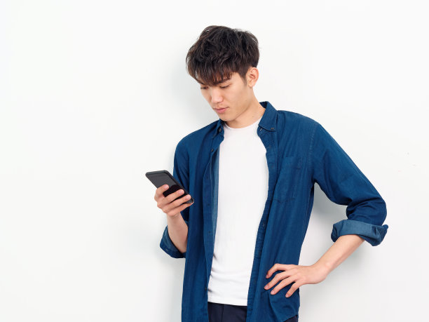 英俊的中国年轻男子的肖像与卷曲的黑色头发在蓝色衬衫摆pose的白墙背景。他手拿智能手机，另一只手放在腰上，拍摄了照片的正面。图片下载