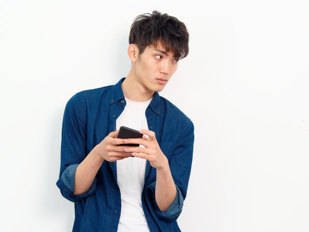 英俊的中国年轻男子的肖像与卷曲的黑色头发在蓝色衬衫摆pose的白墙背景。双手拿着手机看向别处，正面拍摄工作室。图片下载