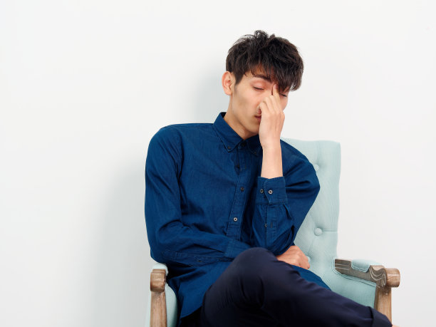 英俊的中国年轻男子的肖像与卷曲的黑色头发，蓝色衬衫坐在扶手椅和摆姿势的白墙背景。手指放在额头上，看起来很累。图片下载