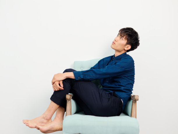 英俊的中国年轻男子的肖像与卷曲的黑色头发，蓝色衬衫坐在扶手椅和摆姿势的白墙背景。双手交叉在膝盖上，全长度侧视图工作室拍摄图片下载