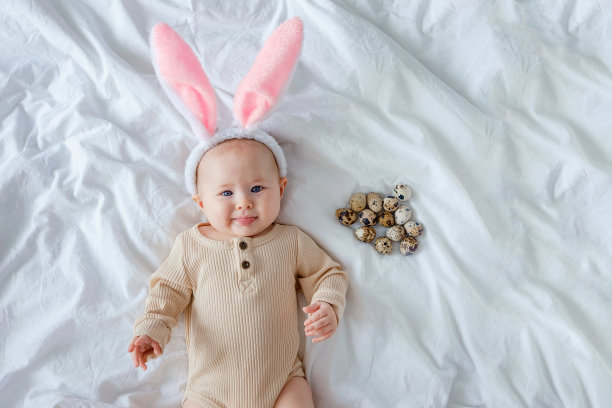 一个可爱的可爱的宝宝穿着复活节的兔子耳朵服装，躺在白色的亚麻布床单上，上面有鹌鹑蛋。一个婴儿看着镜头，露出他的舌头。孩子们的复活节服装。图片下载