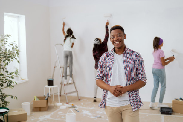 穿着格子衬衫，面带微笑的帅哥正在和他的妻子和女儿们装修一间公寓，女人们正在用白色涂料滚轮为背景粉刷墙壁，装修一间房间图片下载