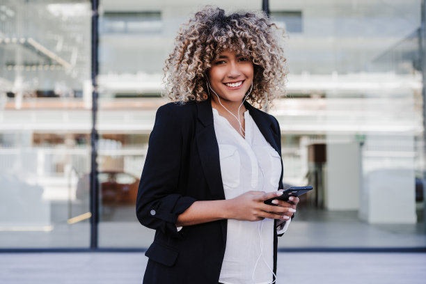 美丽的微笑商业妇女使用手机和耳机在城市。建筑的背景图片下载