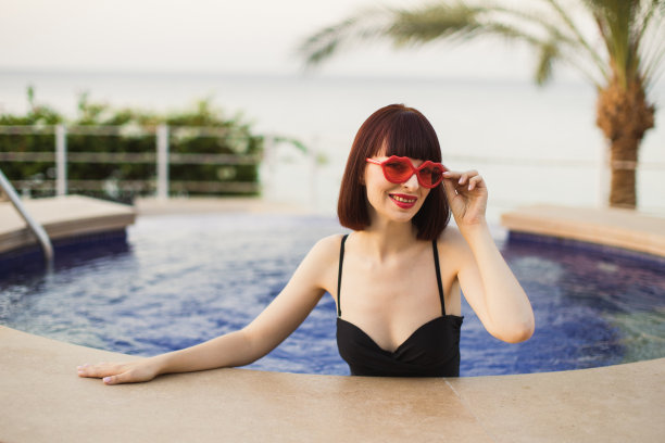 身穿黑色泳衣，戴着红色太阳镜的年轻女性在泳池里享受着阳光和炎热的夏天图片下载