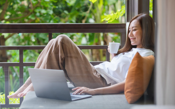 一名年轻女子一边在家里喝咖啡，一边躺在沙发上用笔记本电脑工作或学习图片下载