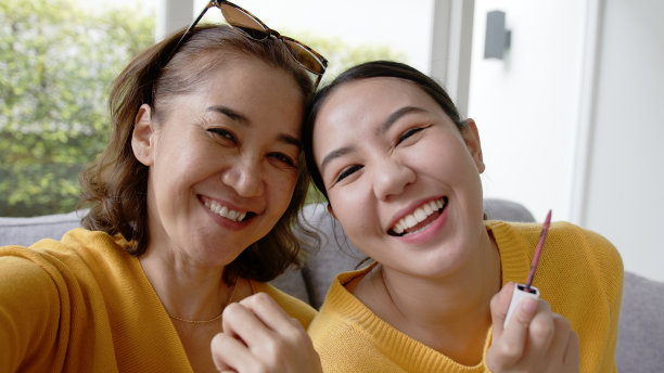 成熟的老妈妈和女孩看着镜头玩有趣的游戏，在亚洲母亲节穿好生活温暖的时候。可爱的青少年喜欢享受社交媒体应用，在家里的沙发上放松。图片下载
