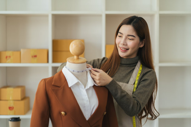侧面的观点，沉思的女裁缝站在测量卷尺附近的人体模型，而缝纫新的时尚服装在现代工作室图片下载