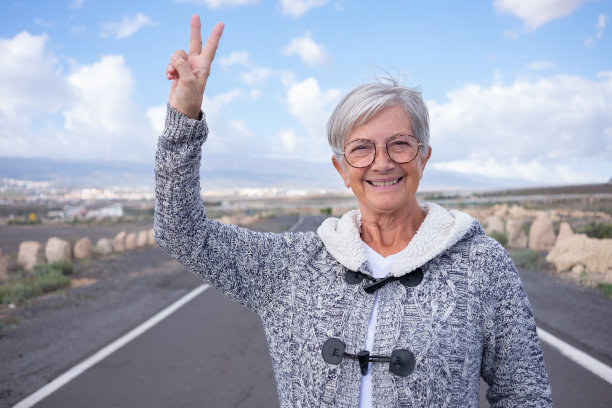 微笑着迷人的年老女子享受户外看着相机手势胜利的手。年长的女士独自走在空旷的路上，背景是山和蓝天图片下载