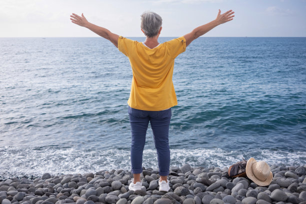 后视镜成熟的老女人，张开双臂迎向大海。白人女性站在卵石海滩享受自由，旅游，度假图片下载