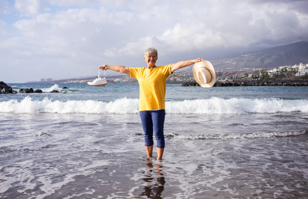 幸福微笑的老年妇女走在海边，帽子和鞋在手中。成熟的女人站在水里，穿着衣服看着镜头。风景优美的自然和背景的地平线图片下载