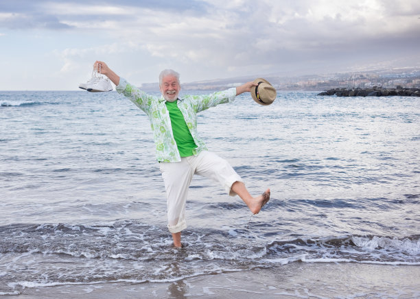 快乐的年老迷人的男子站在海岸线上赤脚，开玩笑地抬起一条腿。成熟的男人手里拿着鞋子和帽子，享受着海滩。轻松快乐的退休老人概念图片下载