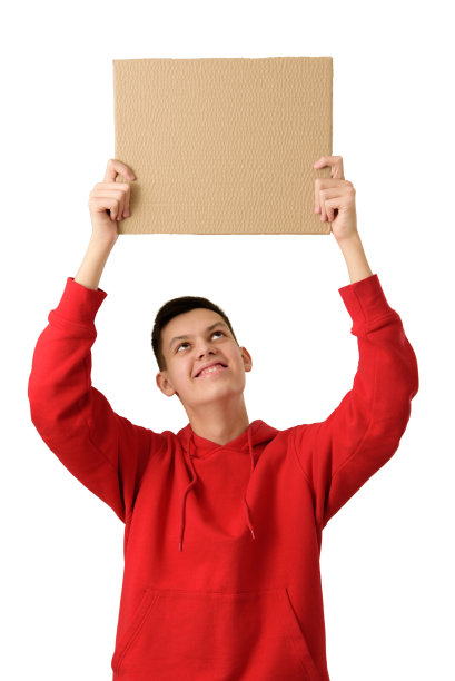 一个十几岁的男孩面带微笑，身穿红色连帽衫，头顶举着空白的棕色纸板横幅，背景是白色的。求职、销售、广告、促销概念。特写镜头。图片下载