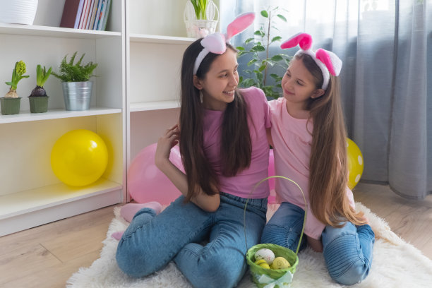 穿着粉红色t恤、耳朵戴着兔子耳朵、面带微笑的姐妹女孩在用气球装饰的房间里收集复活节彩蛋图片下载