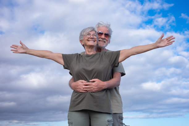 快乐的老男人拥抱他的妻子，而她张开双臂站着。美丽的微笑的白发老人夫妇享受退休和自由的户外旅行图片下载