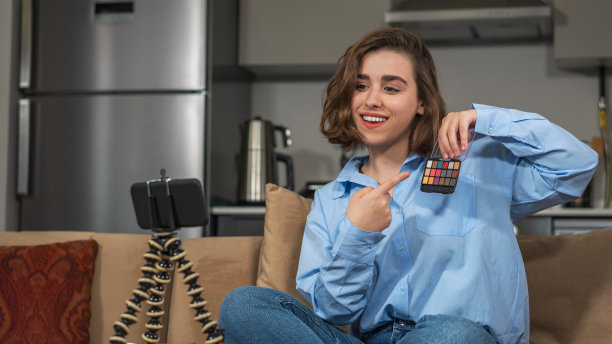 坐在沙发上的小女孩用三脚架接着手机拍照。这个女孩正在展示她手里的化妆用品。年轻的成年人和流行的视频博主坐在家里，用现代手机录制视频。图片下载