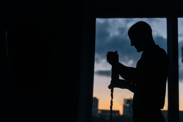 一个无法辨认的肌肉发达的拳击手的剪影，他手腕上缠着拳击胶带，站在傍晚日落时分的窗户背景上。图片下载