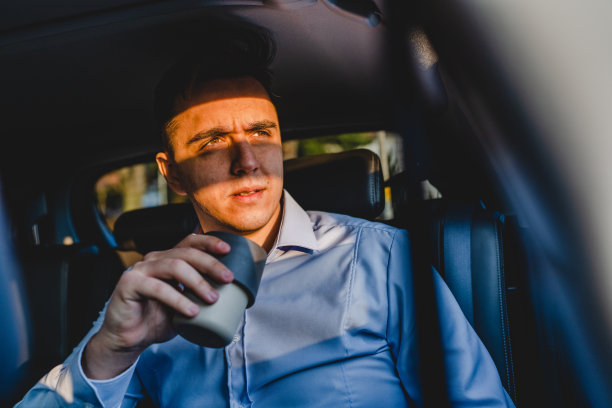 一名男子年轻的成年男性坐在汽车的后座，穿着衬衫，拿着一个咖啡杯或饮料看着旁边的商人经理要去工作的真实的人复制空间图片下载