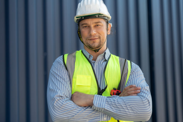 一名体力劳动者身穿绿色工作服，头戴安全帽，自信地站在货仓集装箱前。船舶运输工人概念。图片下载