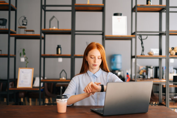 迷人的年轻红发女人的肖像坐在书桌与笔记本电脑在现代的办公室和检查时间的腕表。图片下载