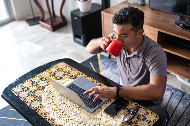 中年亚洲穆斯林男子喝着咖啡，在家里的客厅里工作图片下载